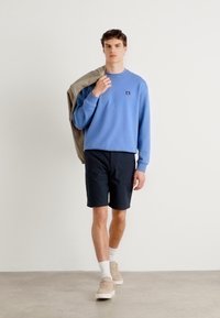 Lys blå sweatshirt med rund hals, liten logo, og svart shorts. Modellen har på seg beige joggesko og hvite sokker.