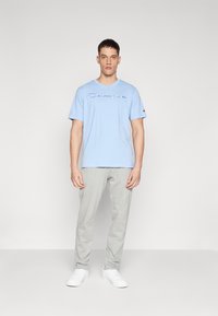 Helles blaues Baumwoll-T-Shirt mit einem weißen "Champion"-Logo, kombiniert mit grauen Sweatpants und weißen Turnschuhen. Einfaches, entspanntes Design.