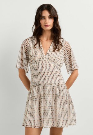 Femme portant une robe courte et fluide avec un motif de points verticaux multicolores et des manches courtes volantes, debout devant un fond clair uni.