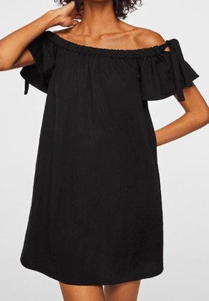 Femme portant une robe noire ample épaules dénudées avec des manches courtes nouées en nœuds, debout avec une main sur la hanche.