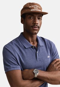 Mann trägt braune Kappe mit weißem Text, blaues Poloshirt und silberne Armbanduhr, Arme verschränkt, neutraler Gesichtsausdruck, hellgrauer Hintergrund.