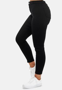 Mujer vestida con jeans negros ajustados y zapatillas blancas, de pie con una rodilla ligeramente doblada contra un fondo claro y liso.