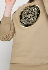 Sudadera beige con un logo bordado redondo en negro y dorado que presenta una abeja, patrones geométricos y el texto "GLORIOUS GANGSTA."