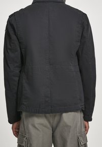 Svart, lätt jacka med strukturerad design, synliga sömmar och krage, kombinerad med grå cargobyxor. Visar baksidan.