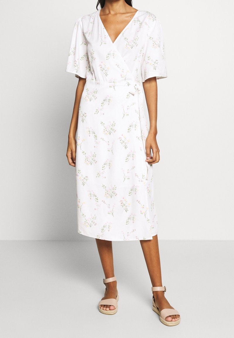 Robe portefeuille blanche à manches courtes, ornée d'un motif floral rose et vert. Longueur mi-mollet avec une ceinture à nouer à la taille.