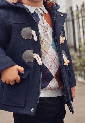 Granatowy wełniany płaszcz z jasnobrązowymi akcentami i zapięciem na toggle. Nałożony na kolorowy sweter w romby i czarne spodnie.