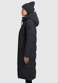 Abrigo de plumas largo y negro con capucha, cierre frontal con cremallera, bolsillos laterales y un parche de marca en la manga. Gorro de punto negro llevado en la cabeza.