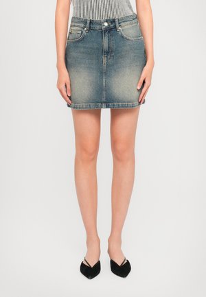 Ženska v izbledelem modrem denim mini krilu s sprednjimi žepi in črnimi čevlji s koničastim nosom, ki stoji pred enotno ozadjem.