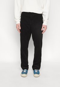 Personne portant un pantalon noir ample, un haut blanc à manches longues et des baskets bleues avec un motif ondulé rouge et noir.