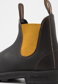 Bota de tornozelo em couro castanho com um painel lateral elástico amarelo, costura de contraste e sola de borracha preta. Logotipo da marca estampado no calcanhar.