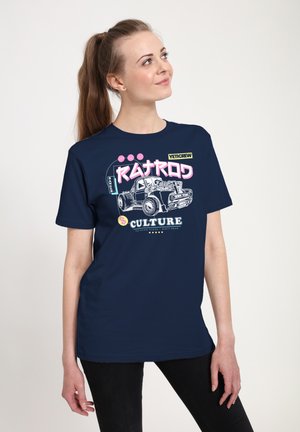 Ung kvinde iført navyblå T-shirt med farverig vintage lastbilgrafik og tekst, der lyder "Railroad Culture", stående mod en ensfarvet baggrund.