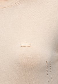 Gros plan sur un tissu tricoté beige avec le logo crocodile tonal Lacoste brodé près de la zone de la poitrine et de petits trous décoratifs sur le côté.