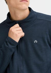 Marineblaue Zip-Jacke mit hohem Kragen, strukturiertem Stoff und weißem Logo auf der linken Brust. Verfügt über schmal geschnittene Ärmel und einen Reißverschluss.