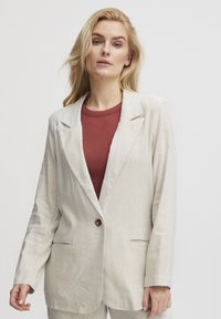 Hellbeige Leinenblazer mit Spitzrevers, einem braunen Knopfverschluss und zwei vorderen Taschen. Darüber wird ein rotes Rundhalsoberteil getragen.