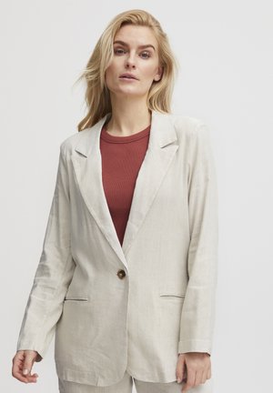 Ljust beige linneblazer med spetsiga slag, en brun knappöppning och två framfickor. Bärs över en röd t-shirt med rund hals.