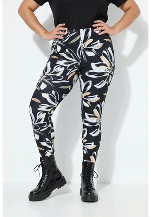 Persoon draagt zwarte gevechtslaarzen en zwarte leggings met grote witte en beige bloemmotieven, handen in de heupen tegen een witte achtergrond.
