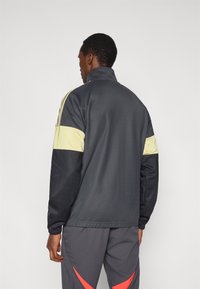 Ľahká športová bunda tmavosivej farby so žltými pruhmi na ramenách. Vysoký golier, dlhé rukávy, hladká textúra a elastické manžety.