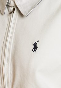 Bílá bavlněná bunda na zip s černým vyšitým logem hráče póla na levé straně a zašpicatělým límcem.