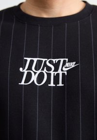 Fekete, rövid ujjú póló függőleges csíkozással. A mellkason fehér hímzett „JUST DO IT” felirat és Nike logó található.