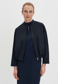 Navy bomber jacket with a round neckline and zip closure, featuring balloon sleeves and smooth texture. Worn over a matching navy dress.