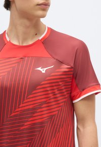 Κοντινό πλάνο ατόμου που φοράει κόκκινο αθλητικό πουκάμισο με γεωμετρικά σχέδια και λευκό λογότυπο Mizuno στο στήθος.