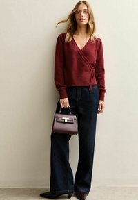 Burgundowa sweter o kroju kopertowym z długimi rękawami i detalem w postaci wiązania, zestawiony z ciemnoniebieskimi spodniami z wysokim stanem i małą burgundową torebką.