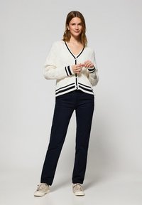Cárdigan blanco de punto trenzado con ribetes y botones de rayas negras, combinado con pantalones navy y zapatillas de color beige claro.