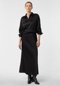 Woman wearing a long black skirt, black button-up shirt with chest pockets, and black ankle boots standing against a plain background.