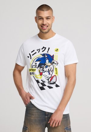 T-shirt bianco in cotone con una grafica stampata di un personaggio riccio blu, con testo colorato e un motivo a scacchi nella parte inferiore.