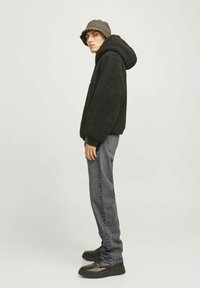 Isik, kes kannab musta fliiskapuutsi, halli kausimütsi, halli teksapükse ja musti kingi, seisab külgvaates tavalise tausta ees.