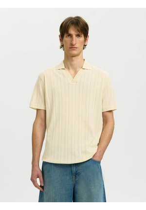 Młody mężczyzna z krótkimi brązowymi włosami, ubrany w kremowy krótki sweter z dzianiny, połączony z luźnymi niebieskimi szortami dżinsowymi, stojący na jednolitym tle.