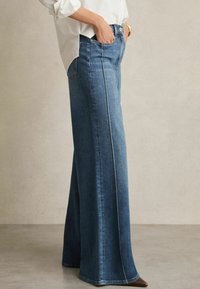 Jeans in denim a gamba larga di colore blu scuro con una texture liscia, vita alta e cuciture a contrasto lungo le suture. Presenta tasche laterali.