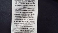 Etichetă de îngrijire a îmbrăcămintei care indică conținutul de țesătură 100% bumbac în mai multe limbi pe o etichetă albă din material textil pe un fundal închis.