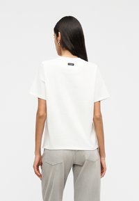 Γυναίκα με μακριά σκούρα μαλλιά που φοράει απλό λευκό μπλουζάκι και ανοιχτό γκρι παντελόνι, με την πλάτη στραμμένη σε λευκό φόντο.