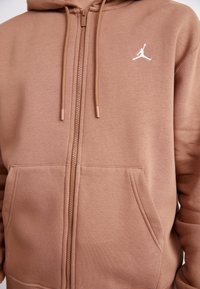 Sudadera con cremallera marrón hecha de un tejido suave, que incluye un bolsillo tipo canguro y un logo Jumpman blanco en el pecho izquierdo. Con cordones acentuados.