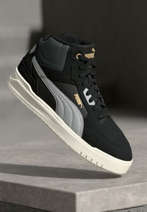 Baskets montantes Puma noires et grises avec une semelle blanche, flottant au-dessus d'une surface en béton sur un fond sombre.