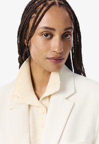 Donna con capelli intrecciati che indossa un maglione a coste color crema sotto a un blazer bianco, guarda leggermente di lato con un'espressione neutra.