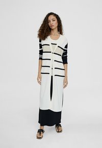 Cardigan lungo con righe orizzontali nere su base bianca, dotato di chiusura a pulsante. Indossato sopra un vestito nero e sandali neri.