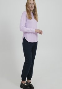 Helllila Pullover mit rundem Halsausschnitt und seitlichen Schlitzen, kombiniert mit navy Jogginghosen. Schwarze Schuhe mit goldfarbener Akzentuierung.