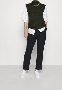 Tmavě zelený žebrovaný pletený svetřík bez rukávů přes bílou košili, v kombinaci s černými střiženými kalhotami a bílými teniskami. Jednoduchý, moderní outfit.