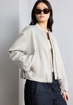 Ženska v bež bomber jakni, temnih kavbojkah in sončnih očalih z okostjem želv, stoji z eno roko v žepu jakne.