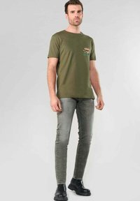 Hombre de pie con camiseta gráfica verde oliva, jeans ajustados grises y botas de cuero negras, contra un fondo claro.