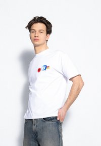 Biała bawełniana koszulka z krótkim rękawem, z kolorowymi nadrukami w czerwonym, niebieskim i żółtym na klatce piersiowej. Nosi w połączeniu z dżinsowymi spodniami.