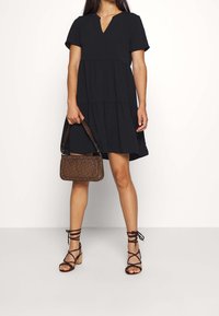 Black tiered dress with short sleeves and a V-neck. Paired with strappy brown heeled sandals and a small patterned handbag.