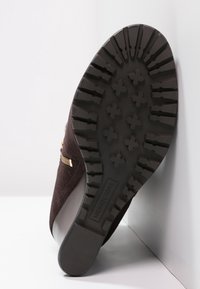 Brun mockaskor med en texturerad svart gummisula med mönstrat mönster på sulan. Guldfärgad metalldetalj på sidan nära en dragkedjedetalj.