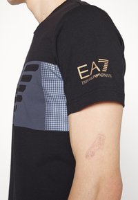 Svart bomullströja med ett grått mönstrat panel och guld EA7-logotyp på ärmen. Enkel design med korta ärmar och rund hals.