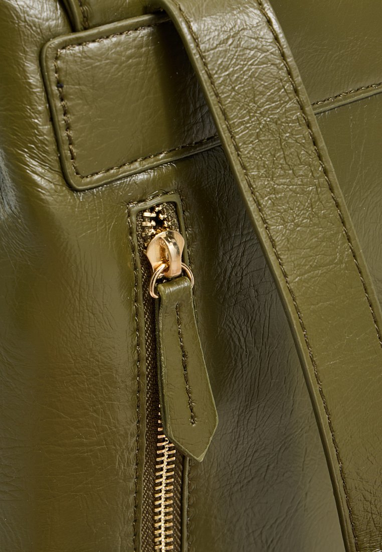 Ecke einer grünen Kunstledertasche mit einem Metallreißverschluss, ausgestattet mit einem goldfarbenen Ziehband und Stichdetails, die eine glatte Textur zeigen.