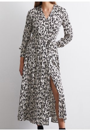 Femme portant une robe portefeuille à manches longues, imprimé animal noir et blanc, avec ceinture et fente latérale.