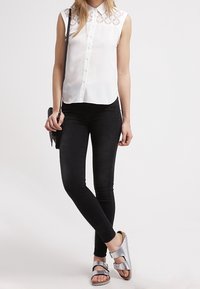 Canottiera bianca senza maniche con yoke in pizzo, jeans skinny neri e sandali argentati con doppie cinghie; il modello tiene una piccola borsa a tracolla nera.