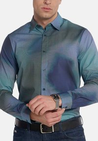 Camicia button-up con un motivo a gradiente blu e teal, maniche lunghe e colletto strutturato. Indossata con una cintura nera e un orologio.